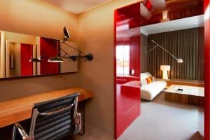 Disfrute de la comodidad de nuestra moderna habitación de hotel con escritorio de madera, silla negra y puerta corredera roja. Relájese en el sofá beige bajo la luz de una gran lámpara. Esta elegante escapada le ofrece el ambiente de una Suite Grand Terrace durante su estancia en Barcelona. - ForTwo - Planes para parejas
