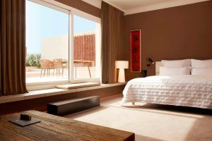 Descubre la Suite Grand Terrace, la comodidad hecha habitación en Barcelona, que cuenta con una amplia estancia con un gran ventanal, cama blanca, mesa de madera y vistas a la terraza con sillas bajo un cielo despejado. - ForTwo - Planes para parejas