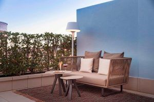 Relájese en nuestra Suite Grand Terrace, la comodidad hecha habitación (Barcelona), con acogedores asientos al aire libre que incluyen un sofá, mesas con bebidas y plantas vibrantes contra una pared azul bajo un cielo despejado. - ForTwo - Planes para parejas