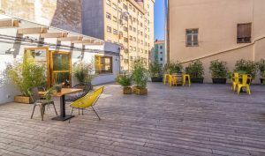 El patio de la azotea del Hostel Bcool Bilbao cuenta con una terraza de madera con sillas y mesas amarillas y negras, y exuberantes plantas en macetas, rodeadas de los imponentes edificios de Bilbao. - ForTwo - Planes para parejas