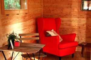 Acogedor espacio de madera en "Habitaciones únicas en Cabañas en los árboles" (Vizcaya) con un sillón rojo vibrante, una pintoresca mesa de madera y vegetación. - ForTwo - Planes para parejas
