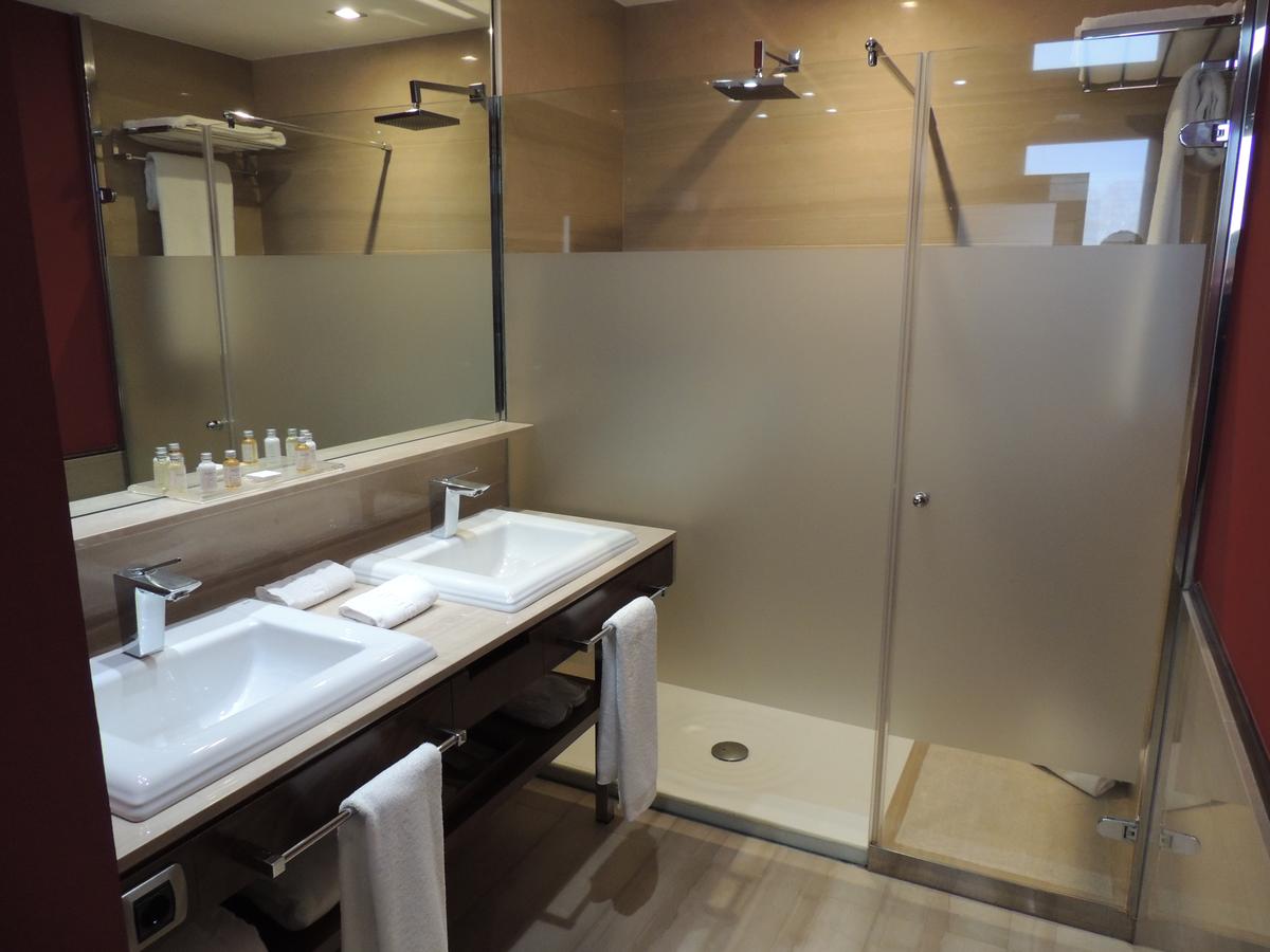 El moderno baño del Hotel Castillo Del Bosque La Zoreda en Oviedo ofrece lavabos dobles, un gran espejo, una cabina de ducha de cristal y lujosas toallas cuidadosamente colocadas en un estante. - ForTwo - Planes para parejas