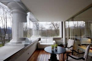 El moderno balcón del Hotel Castillo Del Bosque La Zoreda en Oviedo cuenta con elegantes paredes de cristal, un lujoso sofá blanco y una elegante mesa redonda negra con frondosos árboles que ofrecen un sereno telón de fondo. - ForTwo - Planes para parejas