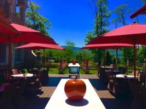 El patio exterior del Hotel Castillo Del Bosque La Zoreda en Oviedo exhibe sombrillas rojas, sillas y esculturas de manzanas en las mesas, todo ello envuelto por una exuberante vegetación bajo el cielo azul claro de la ciudad. - ForTwo - Planes para parejas