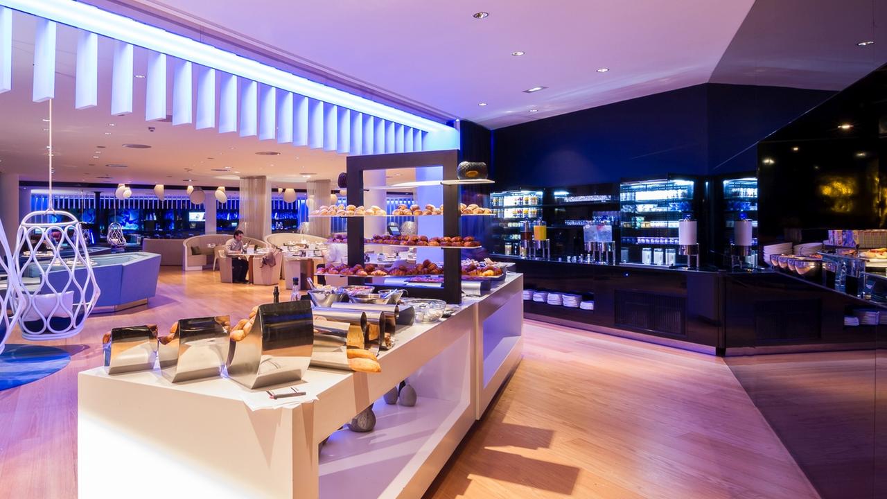 Descubra un moderno buffet en el centro de Barcelona que ofrece repostería y pan, con iluminación azul y blanca y un mobiliario elegante. Su diseño contemporáneo evoca la experiencia Cool Corner Suite del Hotel W: el mar a tus pies. - ForTwo - Planes para parejas