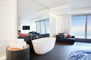 Disfrute del lujo en la Cool Corner Suite: el mar a tus pies en el Hotel W (Barcelona), con un elegante baño con bañera blanca, toallas de felpa y amplios ventanales con impresionantes vistas de la ciudad. Relájese en el sofá junto al elegante espejo de pared para una experiencia inolvidable. - ForTwo - Planes para parejas