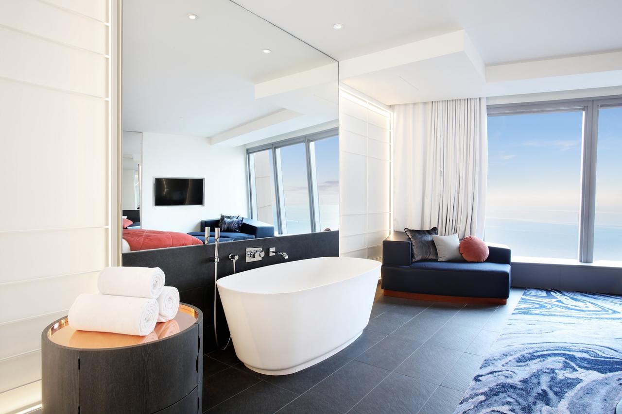 Disfrute del lujo en la Cool Corner Suite: el mar a tus pies en el Hotel W (Barcelona), con un elegante baño con bañera blanca, toallas de felpa y amplios ventanales con impresionantes vistas de la ciudad. Relájese en el sofá junto al elegante espejo de pared para una experiencia inolvidable. - ForTwo - Planes para parejas