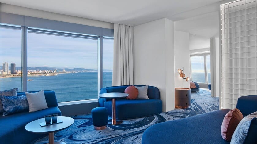 Descubra la Cool Corner Suite: el mar a tus pies en el Hotel W (Barcelona), donde el lujo moderno se fusiona con la modernidad. Esta exquisita habitación cuenta con sofás azules, una alfombra artística y amplios ventanales que ofrecen impresionantes vistas de la costa. - ForTwo - Planes para parejas