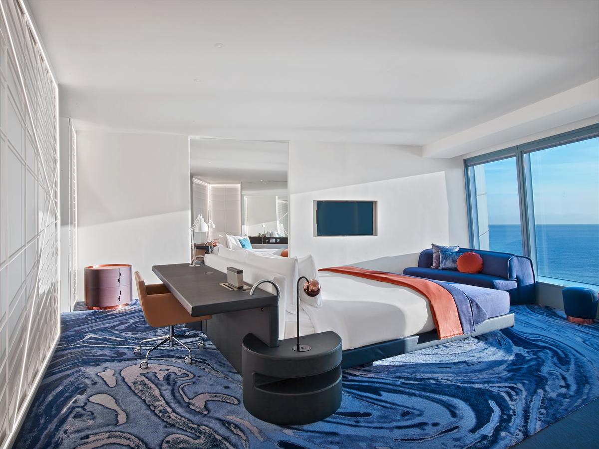La Suite Cool Corner del Hotel W Barcelona ofrece vistas al mar con cama, escritorio, sofá azul y una alfombra azul abstracta. Disfruta del mar a tus pies en esta impresionante habitación. - ForTwo - Planes para parejas