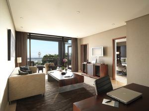 La Gran Suite Mediterránea con terraza del Hotel Miramar Barcelona ofrece una moderna sala de estar con un sofá beige y elegantes muebles de madera. Unas puertas de cristal dan a una terraza con impresionantes vistas al mar, mientras que un escritorio alberga un portátil, combinando a la perfección lujo y tecnología. - ForTwo - Planes para parejas