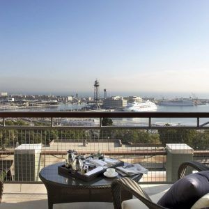 Deléitese con la extraordinaria vista desde la Gran Suite Mediterránea con terraza en el Hotel Miramar (Barcelona), que ofrece una perspectiva perfecta del puerto, sus majestuosos cruceros y un paisaje urbano de fondo, todo ello realzado por una acogedora mesa preparada con un encantador servicio de té. - ForTwo - Planes para parejas