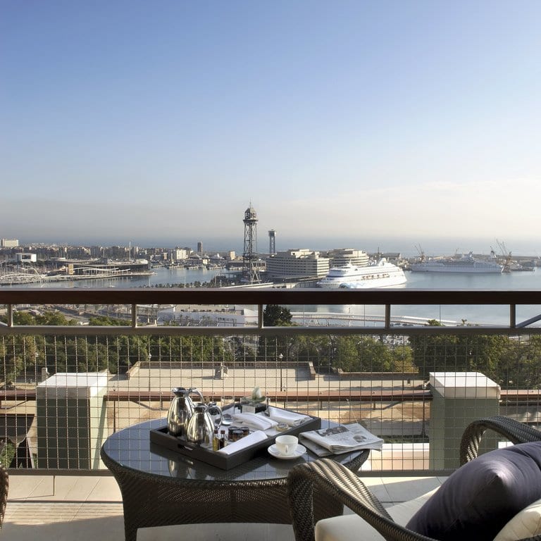 Deléitese con la extraordinaria vista desde la Gran Suite Mediterránea con terraza en el Hotel Miramar (Barcelona), que ofrece una perspectiva perfecta del puerto, sus majestuosos cruceros y un paisaje urbano de fondo, todo ello realzado por una acogedora mesa preparada con un encantador servicio de té. - ForTwo - Planes para parejas