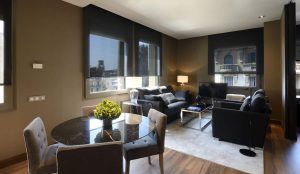 Una moderna sala de estar en Barcelona con sofás oscuros, TV y grandes ventanales similar a la Grand Suite: la joya de la corona del Grand Hotel Central (Barcelona), con una mesa de comedor y sillas sobre un hermoso piso de madera. - ForTwo - Planes para parejas