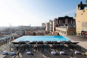 Disfrute de la piscina infinita en la azotea de la Grand Suite, la joya de la corona del Grand Hotel Central, con tumbonas alineadas que ofrecen impresionantes vistas del paisaje urbano de Barcelona bajo un cielo azul claro. - ForTwo - Planes para parejas