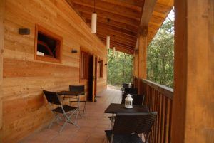 Un porche de madera con sillas y mesas, rodeado de árboles, refleja la serenidad de la Habitación mágica en los árboles por Cabanes Dosrius. Las luces colgantes y las linternas crean un ambiente acogedor en esta tranquila escapada. - ForTwo - Planes para parejas