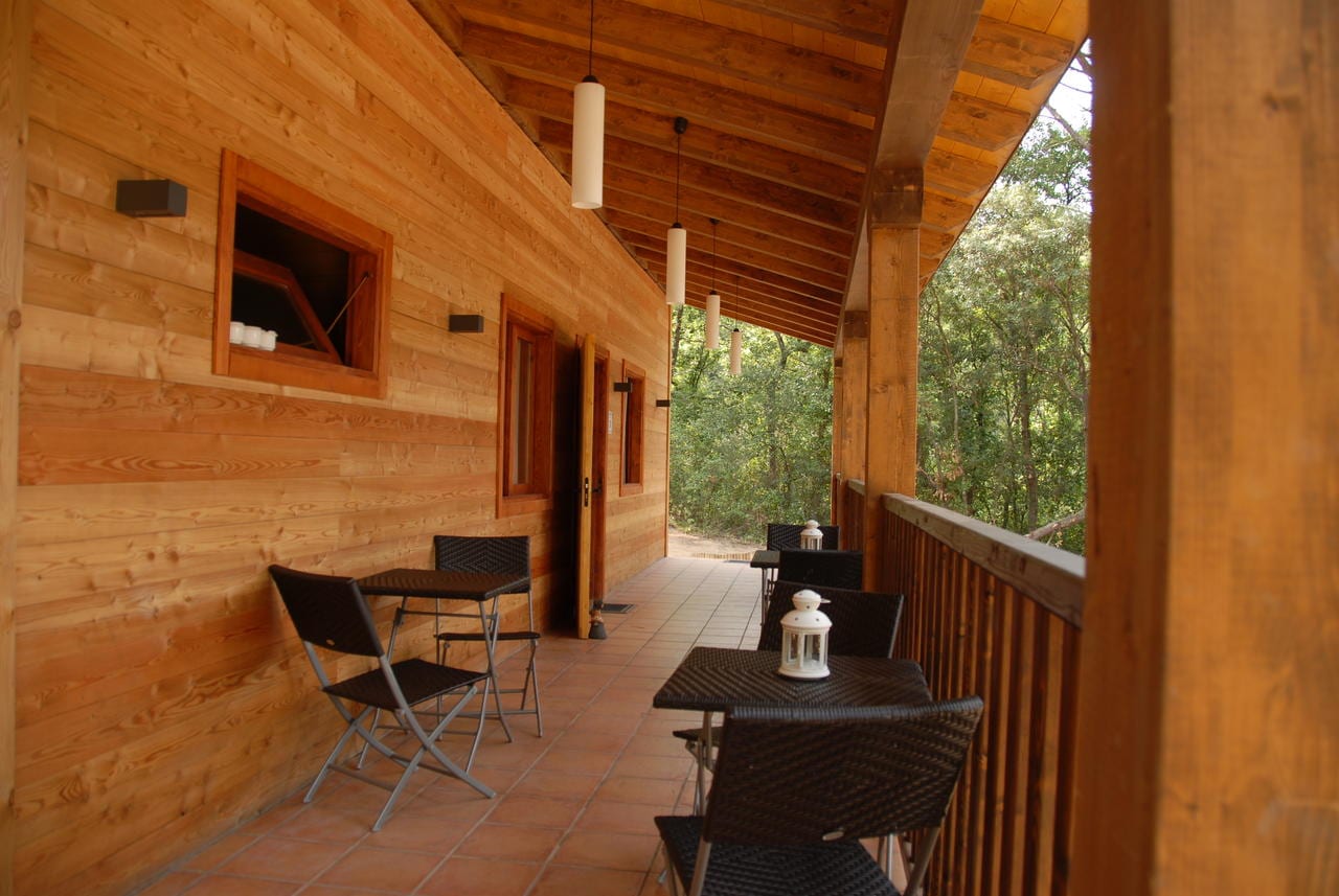 Un porche de madera con sillas y mesas, rodeado de árboles, refleja la serenidad de la Habitación mágica en los árboles por Cabanes Dosrius. Las luces colgantes y las linternas crean un ambiente acogedor en esta tranquila escapada. - ForTwo - Planes para parejas