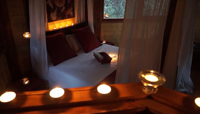 Un acogedor dormitorio cuenta con una cama con dosel, iluminación tenue y velas. Cortinas blancas y cojines rojos evocan el ambiente romántico de una Habitación mágica en los árboles por Cabanes Dosrius en Barcelona. - ForTwo - Planes para parejas