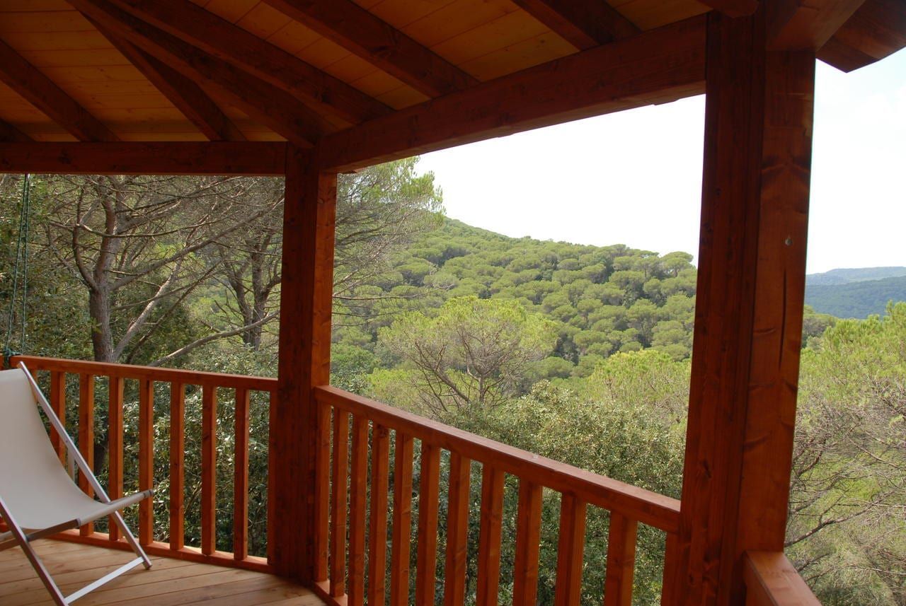 Una silla blanca sobre un porche de madera da a un frondoso bosque verde, evocando la encantadora esencia de la Habitación mágica en los árboles por Cabanes Dosrius (Barcelona). - ForTwo - Planes para parejas