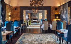 El elegante restaurante del Hotel Castell d'Emporda, Girona, luce paredes de piedra, lámparas de araña y lujosas sillas azules. Las mesas cuentan con servilletas y vajilla blancas para crear un ambiente sofisticado, ideal para cualquier experiencia gastronómica. - ForTwo - Planes para parejas