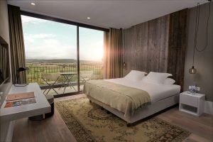 Una acogedora habitación en el Hotel Castell d'Emporda (Girona) incluye una cama grande, una pared con detalles de madera y un balcón con vistas al pintoresco paisaje del atardecer de Girona. - ForTwo - Planes para parejas