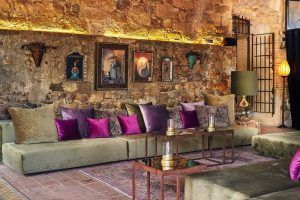 Una habitación rústica del Hotel Castell d'Emporda en Girona exhibe paredes de piedra, sofás de terciopelo verde, almohadas moradas, pinturas exquisitas y una iluminación cálida. - ForTwo - Planes para parejas