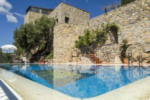 El Hotel Castillo de Monda (Málaga), un edificio de piedra, está enclavado entre frondosos árboles con una piscina en primer plano bajo el cielo azul parcialmente nublado. - ForTwo - Planes para parejas