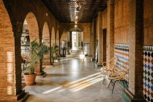 En el soleado pasillo del Hotel Castillo de Monda (Málaga), paredes de ladrillo arqueadas, decoración de azulejos y sillas de mimbre se combinan armoniosamente, con plantas que bordean el camino, capturando el vibrante encanto de Málaga en cada rincón. - ForTwo - Planes para parejas