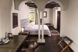 El Hotel Castillo de Monda en Málaga ofrece un acogedor dormitorio con una cama doble, muebles de madera oscura, azulejos decorativos en la pared, una entrada arqueada y una ventana con vistas panorámicas a los pintorescos paisajes. - ForTwo - Planes para parejas