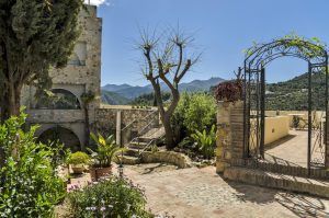 El Hotel Castillo de Monda (Málaga) cuenta con un patio de piedra con exuberante vegetación y una puerta de hierro forjado que se abre a las vistas a la montaña bajo un cielo azul, combinando el encanto rústico con la belleza natural. - ForTwo - Planes para parejas
