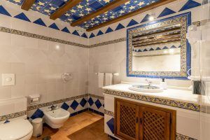 Este encantador baño refleja la elegancia del Hotel Castillo de Monda de Málaga, con un patrón de azulejos azules y blancos, un techo con vigas de madera y un espejo ornamentado sobre el lavabo. - ForTwo - Planes para parejas