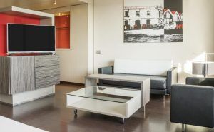 Una moderna sala de estar, que recuerda a la Junior Suite: el secreto de Ilunion Hotels en Barcelona, exhibe un televisor de pantalla plana, un sofá gris, una mesa de café con tapa de cristal y llamativas obras de arte en blanco y negro. - ForTwo - Planes para parejas