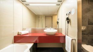 Experimente la elegancia y la comodidad en la Junior Suite de El Secreto de Ilunion Hotels, Barcelona. Cuenta con un moderno baño con una elegante encimera roja, un lavabo blanco impecable, un gran espejo, una bañera y un toallero. - ForTwo - Planes para parejas