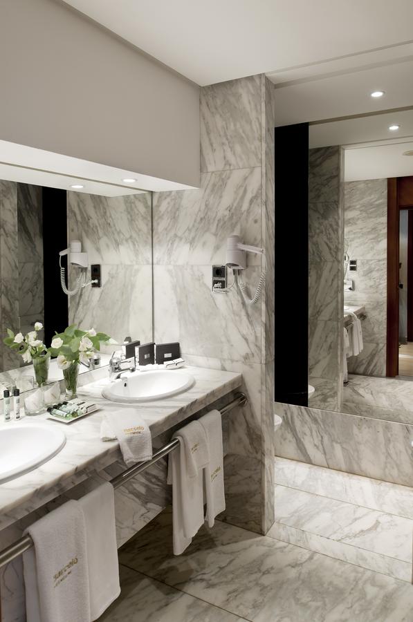 La suite presidencial del Hotel Barceló Sevilla Renacimiento cuenta con un elegante baño de mármol con lavabos dobles, espejos de pared, toallas blancas y flores frescas. - ForTwo - Planes para parejas