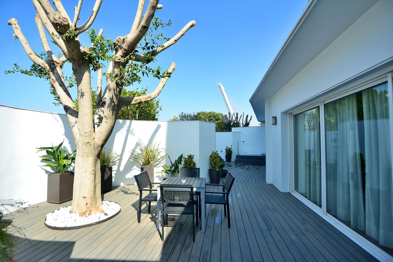 Descubra la sofisticada terraza en la azotea de La Suite Presidencial del Hotel Barceló Sevilla Renacimiento en Sevilla. Una elegante terraza de madera, una mesa de cristal, sillas elegantes, un árbol desnudo y plantas vibrantes complementan las paredes blancas y el cielo despejado para un estilo inigualable. - ForTwo - Planes para parejas