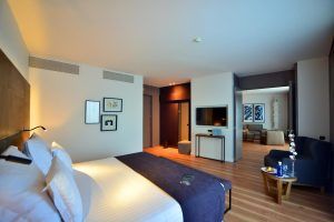 Descubra la elegancia de La Suite Presidencial del Hotel Barceló Sevilla Renacimiento, con una cama grande, impactantes obras de arte en las paredes, entretenimiento con TV y una acogedora sala de estar con elegantes suelos de madera. Los tonos blancos y oscuros se combinan a la perfección para crear un ambiente sofisticado. - ForTwo - Planes para parejas