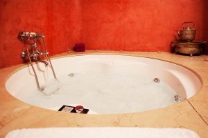 Alójese en una suite romántica en Alcoba del Rey de Sevilla, que ofrece una bañera redonda con chorros de jacuzzi, rodeada de paredes de estuco rojo y realzada por una tetera de metal vintage sobre un soporte ornamentado. - ForTwo - Planes para parejas