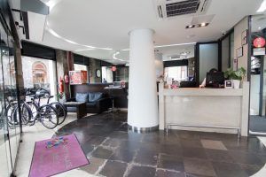 El moderno vestíbulo del Hotel Petit Palace luce elegantes sofás negros y bicicletas junto a la ventana, creando un ambiente único. Tras el mostrador de recepción, un miembro del personal le espera para garantizar su estancia de ensueño en el Hotel Petit Palace (Bilbao). - ForTwo - Planes para parejas