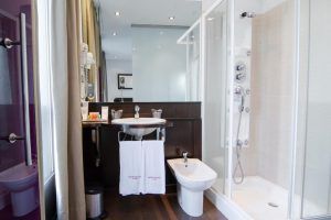 Disfrute de un baño moderno en la Estancia de Ensueño del Hotel Petit Palace Bilbao, con ducha de cristal, lavabo con espejo, bidé y toallas de lujo. Disfrute de una decoración minimalista y suelos de madera para una estancia de ensueño en el corazón de Bilbao. - ForTwo - Planes para parejas
