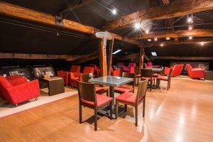 La Estancia de ensueño en Hotel Petit Palace (Bilbao) cuenta con un acogedor salón abuhardillado con vigas de madera, sillones rojos y sillas marrones sobre suelo de madera. - ForTwo - Planes para parejas