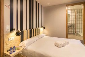Descubre el elegante alojamiento del Hostel Bcool Bilbao, con un diseño moderno, una cama doble, una pared con acento de rayas, toallas enrolladas, botellas de agua y una vista única al baño, todo en el vibrante corazón de Bilbao. - ForTwo - Planes para parejas