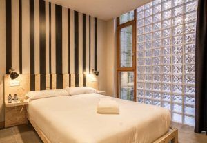La moderna habitación del Hostel Bcool Bilbao cuenta con una pared con detalles en blanco y negro a rayas, una ventana de bloques de vidrio y cuenta con una cama cuidadosamente hecha y toallas incluidas. - ForTwo - Planes para parejas