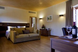 Elegante habitación en la Estancia en la Abadía Retuerta LeDomaine, Valladolid, con cama doble, sofá, mesa con la botella de vino LeDomaine, televisor y un gran ventanal con cortinas. La cálida iluminación realza el ambiente acogedor. - ForTwo - Planes para parejas