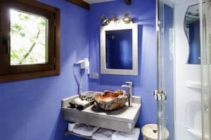 Este moderno baño, que refleja el estilo elegante de la Habitación Casa del árbol en el Hotel Maitenea Jakue en Navarra, cuenta con una llamativa pared azul, detalles de madera, un lavabo en forma de concha y una elegante ducha de cristal. - ForTwo - Planes para parejas