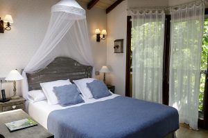 Una acogedora habitación en la Habitación Casa del árbol Maitenea del Hotel Jakue (Navarra) cuenta con una cama con dosel, ropa de cama azul y lámparas de noche. Cortinas transparentes enmarcan una ventana, ofreciendo una serena vista al jardín que evoca el entorno que le da nombre. - ForTwo - Planes para parejas