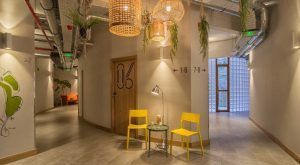 El pasillo del Hostel Bcool Bilbao en Bilbao muestra sillas amarillas, una pequeña mesa y lámparas de mimbre colgantes; los números de las habitaciones se muestran en las paredes. - ForTwo - Planes para parejas