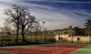 Una serena cancha de tenis con superficie roja descansa en medio del paisaje montañoso de La Posada de Pradorey, rodeada de árboles sin hojas bajo un vasto cielo azul. - ForTwo - Planes para parejas