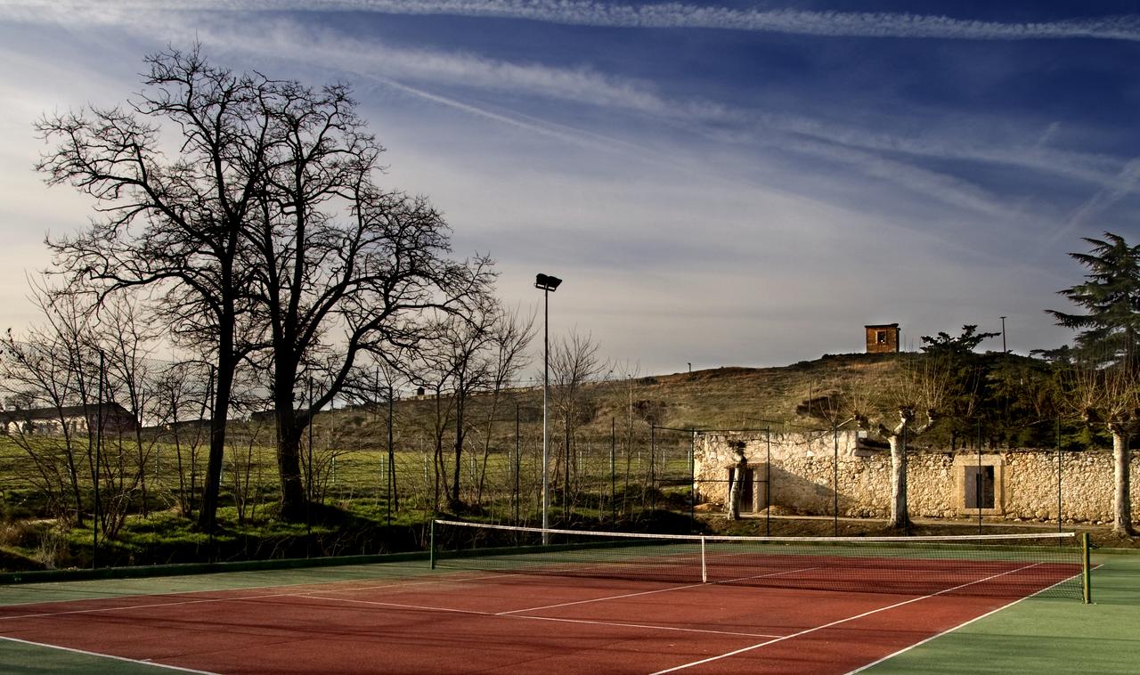 Una serena cancha de tenis con superficie roja descansa en medio del paisaje montañoso de La Posada de Pradorey, rodeada de árboles sin hojas bajo un vasto cielo azul. - ForTwo - Planes para parejas