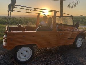 Una pareja disfruta de una vista del atardecer en un descapotable naranja en un camino de grava junto a un campo pintoresco, con viñedos ondulados, capturando la esencia aventurera y refinada del "Día romántico de vino y gastronomía entre viñedos (Barcelona). - ForTwo - Planes para parejas