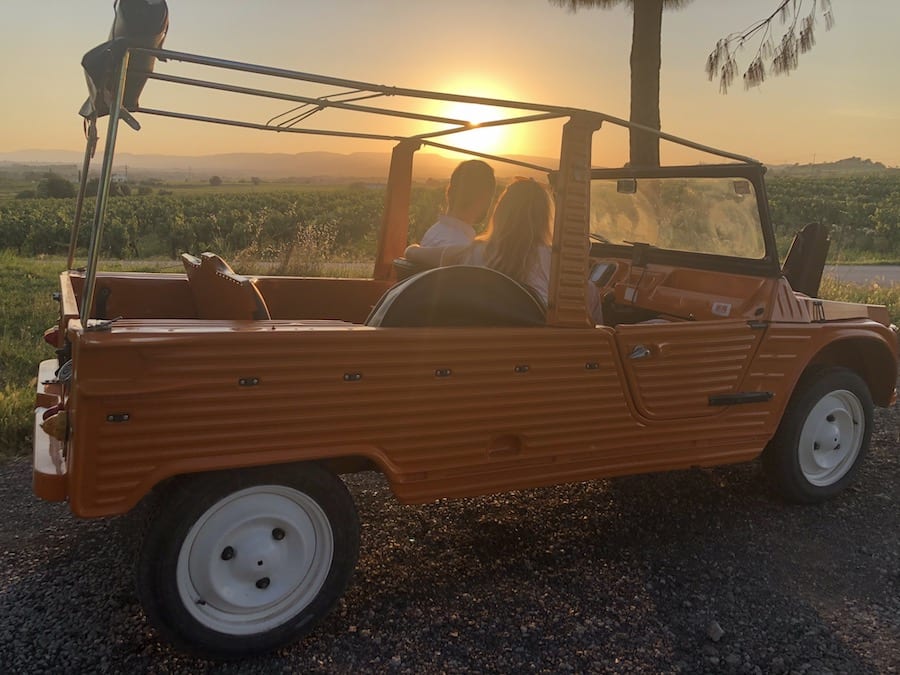 Una pareja disfruta de una vista del atardecer en un descapotable naranja en un camino de grava junto a un campo pintoresco, con viñedos ondulados, capturando la esencia aventurera y refinada del "Día romántico de vino y gastronomía entre viñedos (Barcelona). - ForTwo - Planes para parejas