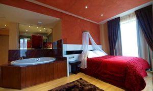 Embárcate en una escapada romántica con el paquete "Escapada romántica en el Hotel Spa Felisa (Santander)". Relájate en una acogedora habitación con colcha roja, jacuzzi, cortinas marrones e iluminación cálida. - ForTwo - Planes para parejas